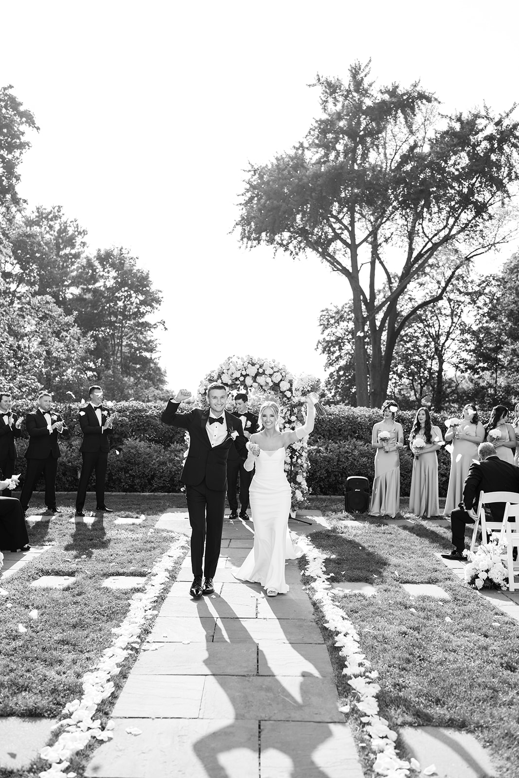 Black and white photo of the newlyweds walking up the aisle, celebrating with raised hands.
